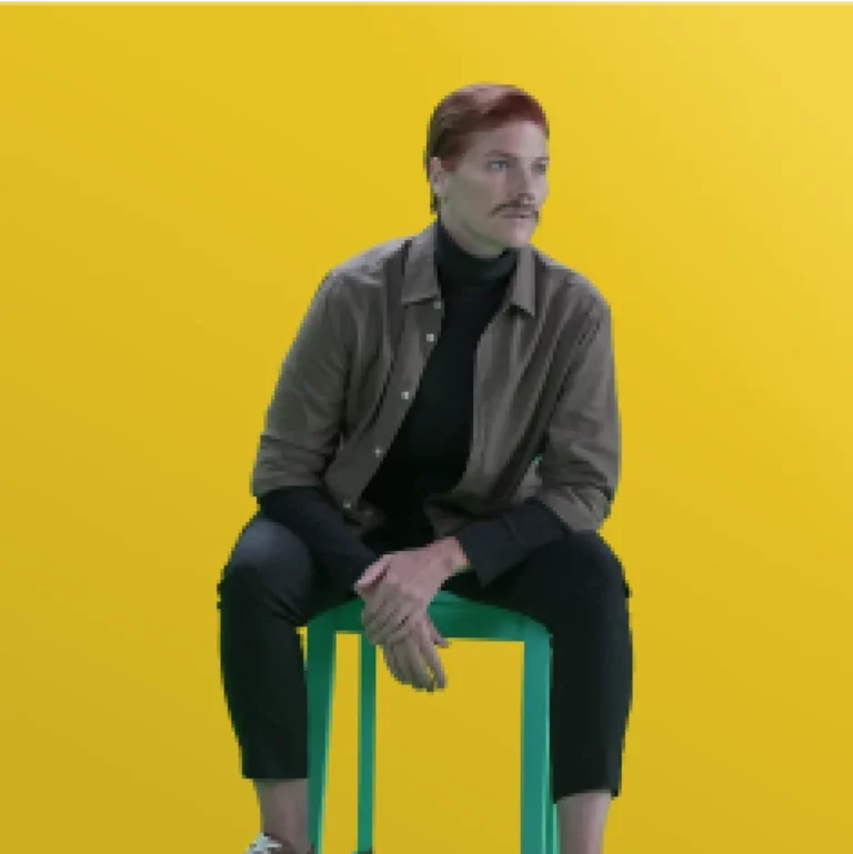 Major Kami poses on a chair with a yellow background, the picture is edited with pixelisation effect